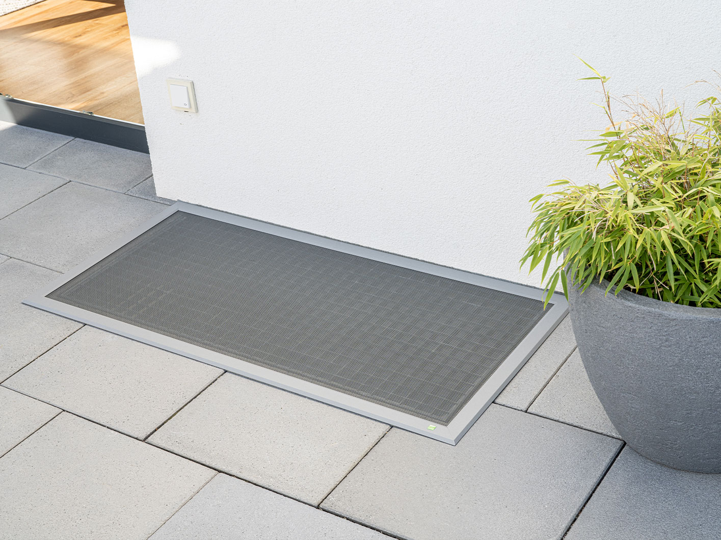 Eine edle Lichtschachtabdeckung auf einer Terrasse. Rechts davon ist eine Tür und links davon eine Topf mit einer Pflanze.